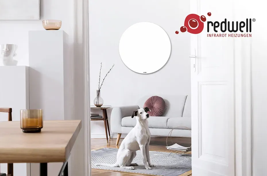 Runde, weiße Redwell Infrarotheizung an der Wand in einem hellen Wohnzimmer mit Sofa und einem sitzenden Hund.