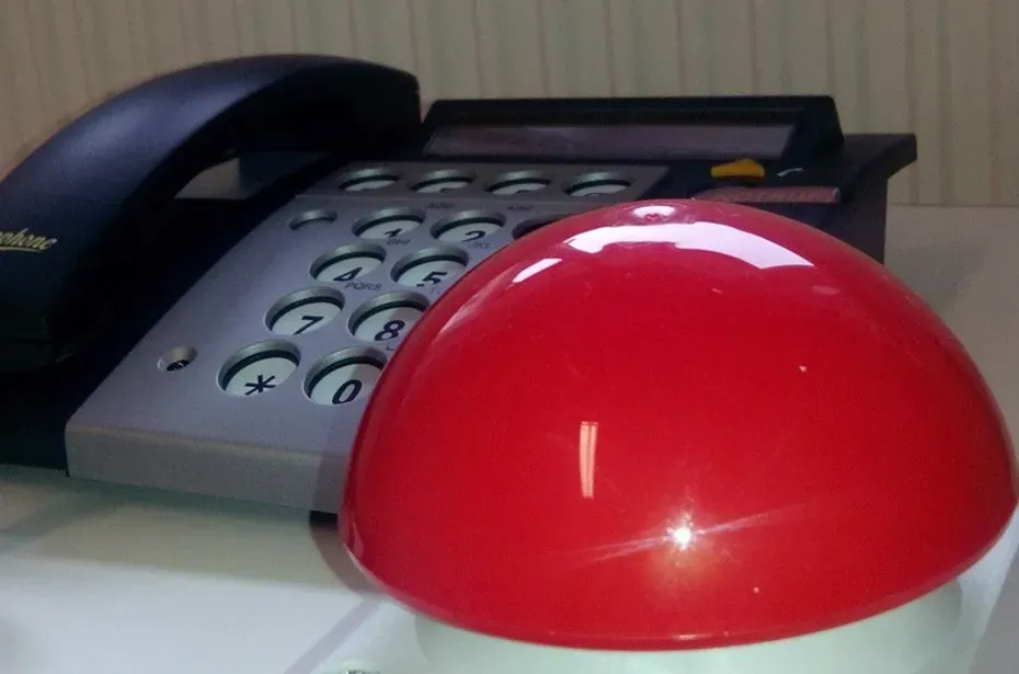 Ein großer, glänzender roter Druckknopf im Vordergrund, dahinter steht ein graues Festnetztelefon mit Hörer.